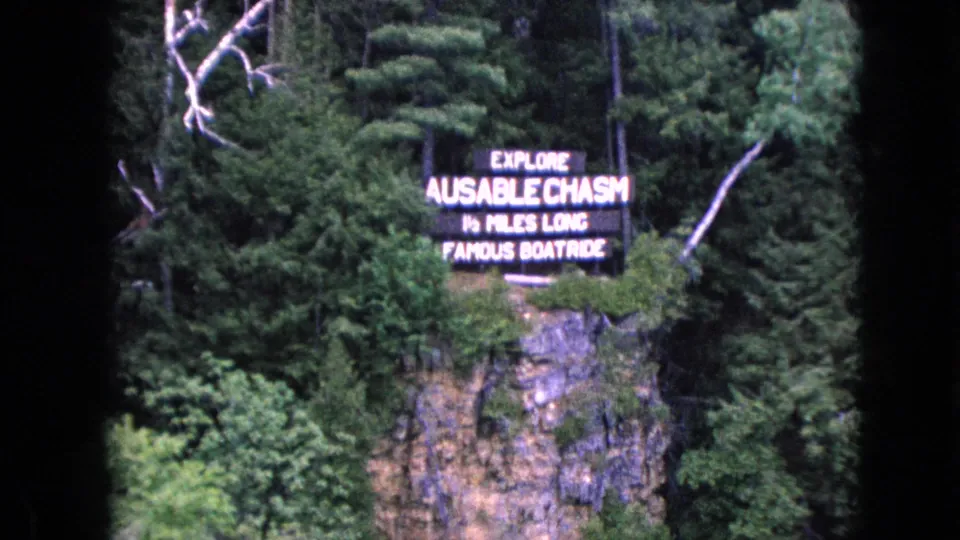 The Ausable Chasm sign appears near the upper right of the frame above a rugged cliff and dense green trees in archival 8mm footage.