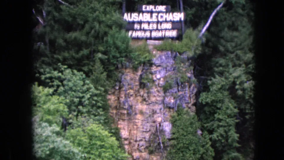 A soft 8mm frame centers the Ausable Chasm tourist sign above a rocky cliff, surrounded by green forest and visible film gate edges.