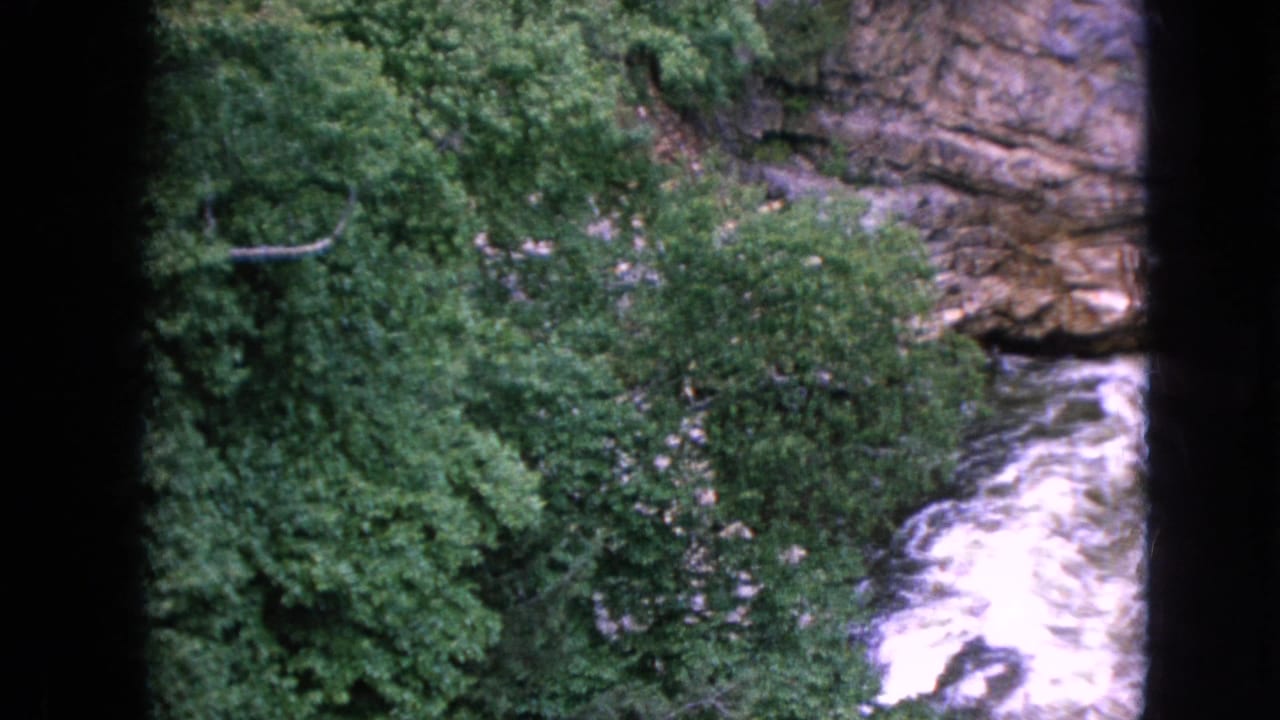 1964 Niagara Falls, New York travel footage frame showing whitewater rapids beside dense green foliage and exposed rock layers in a gorge.