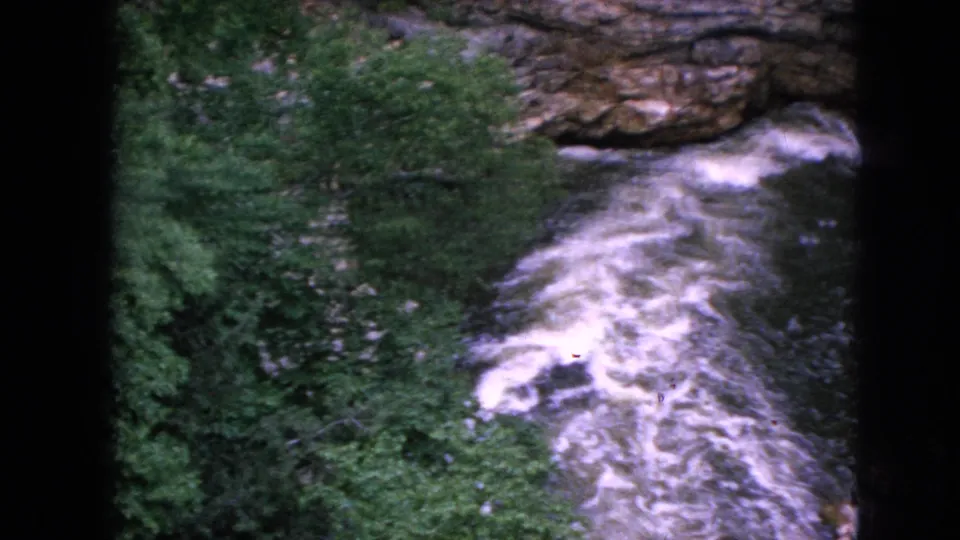 1964 8mm frame of a Niagara Falls area gorge with foaming river water below dense trees and a rough rock ledge, seen from above.