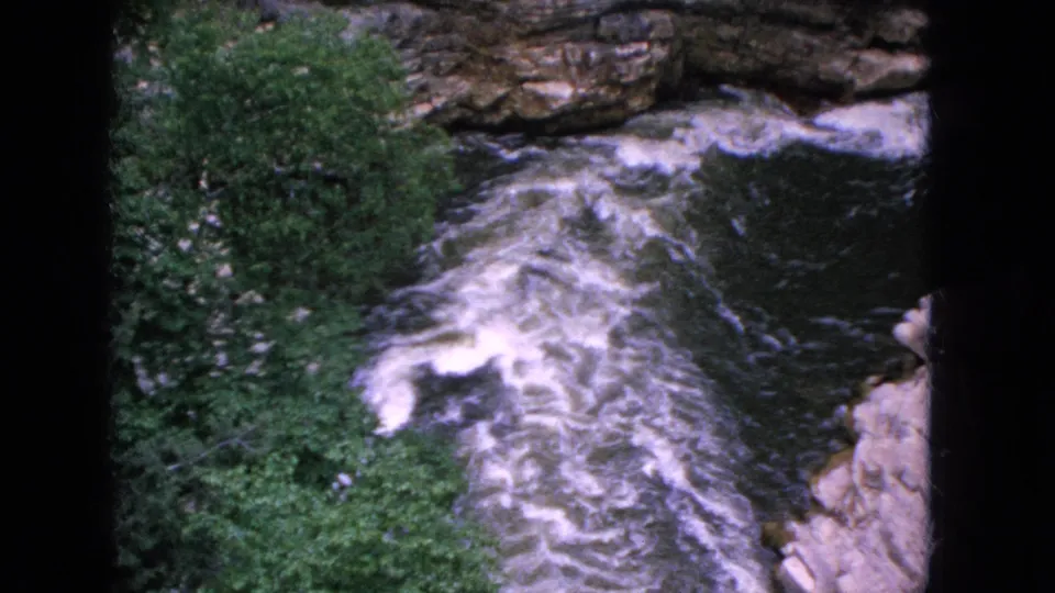 1964 Niagara Falls, New York home-movie frame showing churning rapids in a narrow rocky channel with dense vegetation along the gorge walls.