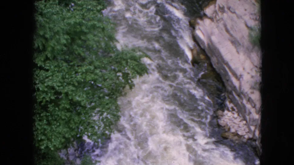 1964 Niagara Falls, New York home-movie still of rushing white water descending through a narrow rock-lined channel beside dense trees.