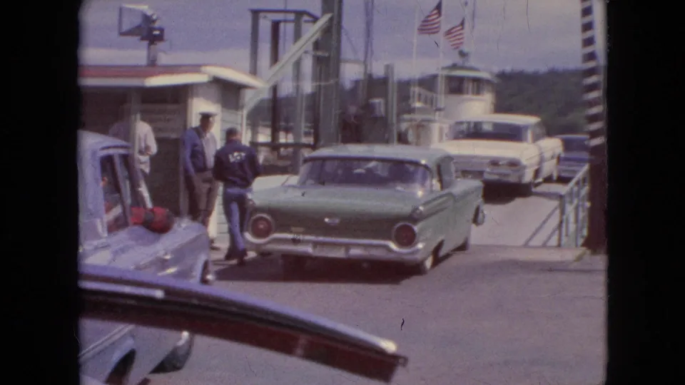 At a Niagara Falls area ferry or dock entrance, mid-century cars queue as several men stand near the gate and American flags fly behind them.