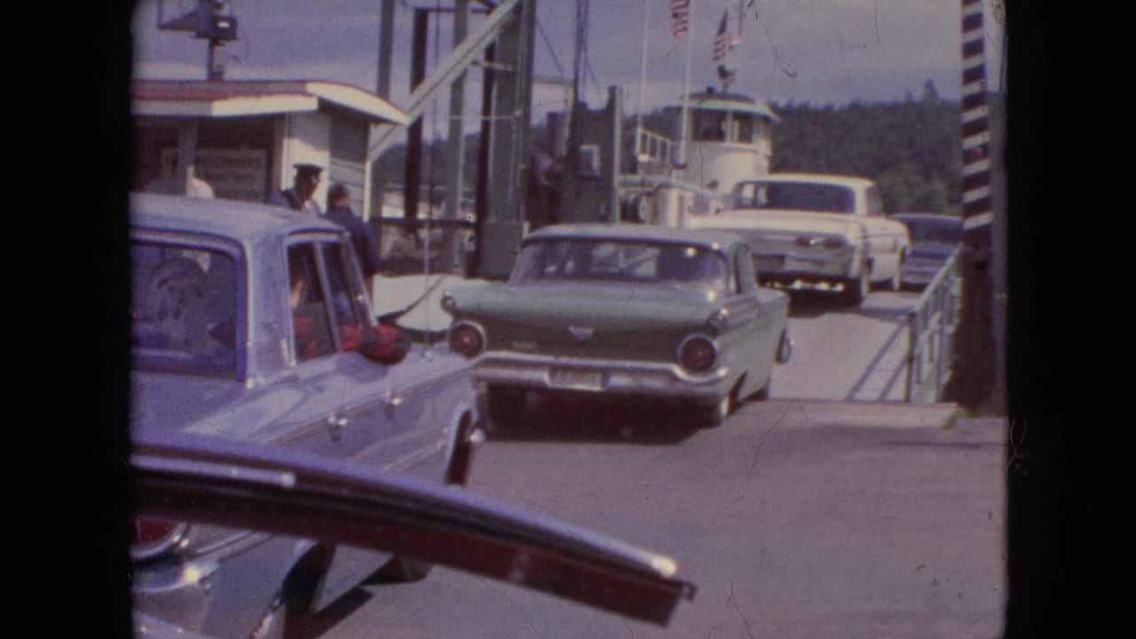 1964 home-movie frame from Niagara Falls, New York, showing mid-century cars waiting on a ferry ramp with attendants and the ferry structure visible.