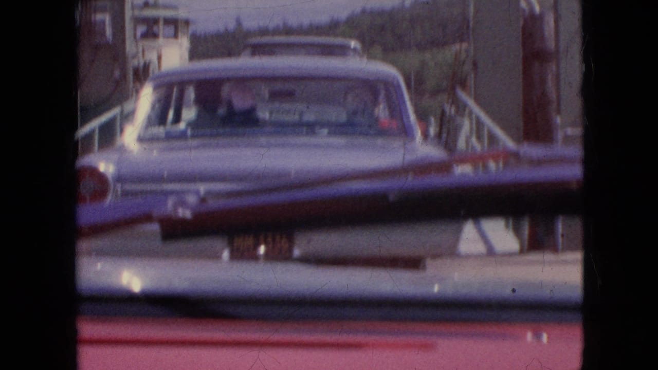 A mid-century sedan ahead on the ferry ramp is seen from inside another car, with the windshield wiper and dashboard edge framing the view.