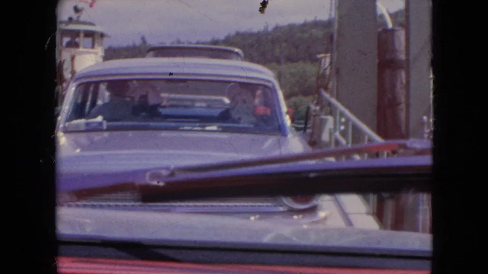 A sedan faces the camera on the ferry ramp, with passengers visible through the windshield and the filming car's wiper crossing the foreground.