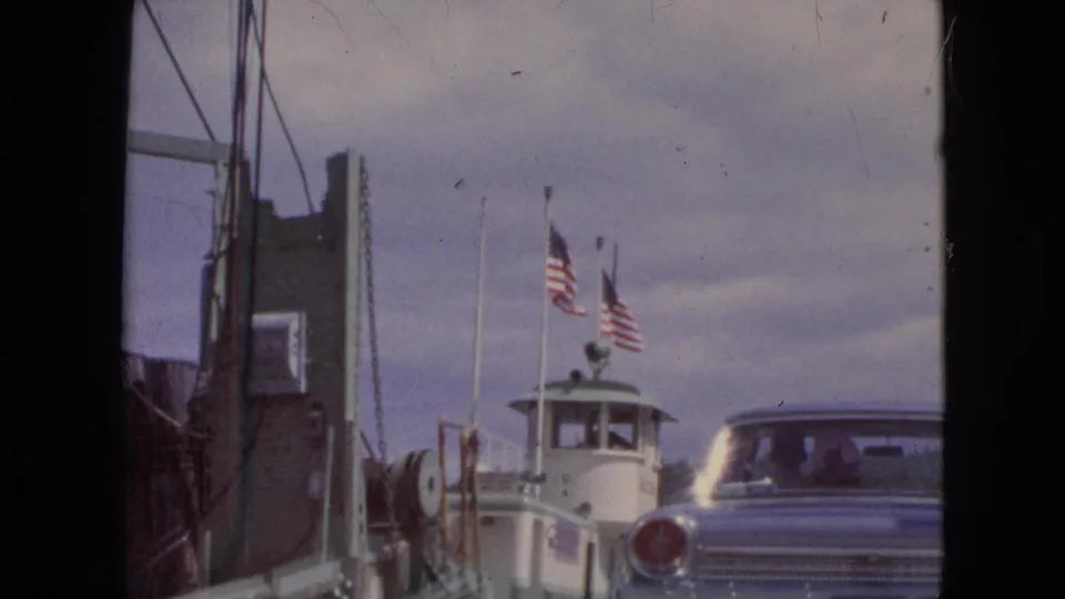 The ferry pilothouse and American flags rise above the ramp as a classic car appears at the right edge in this 1964 travel scene.
