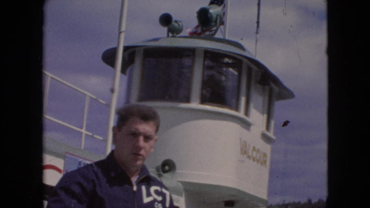 A young man in a navy LCT Co. jacket stands on the deck of the white Valcour ferry, with the pilothouse, loudspeaker, railing, and cloudy sky behind him.