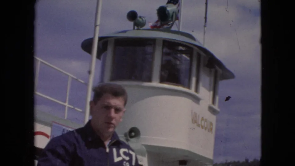 A young man in a navy LCT Co. jacket stands on the deck of the white Valcour ferry, with the pilothouse, loudspeaker, railing, and cloudy sky behind him.