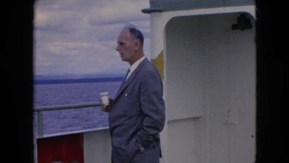 An older man in a gray suit holds a small cup while standing on the ferry deck, with a railing, open water, distant shoreline, and white boat structure around him.