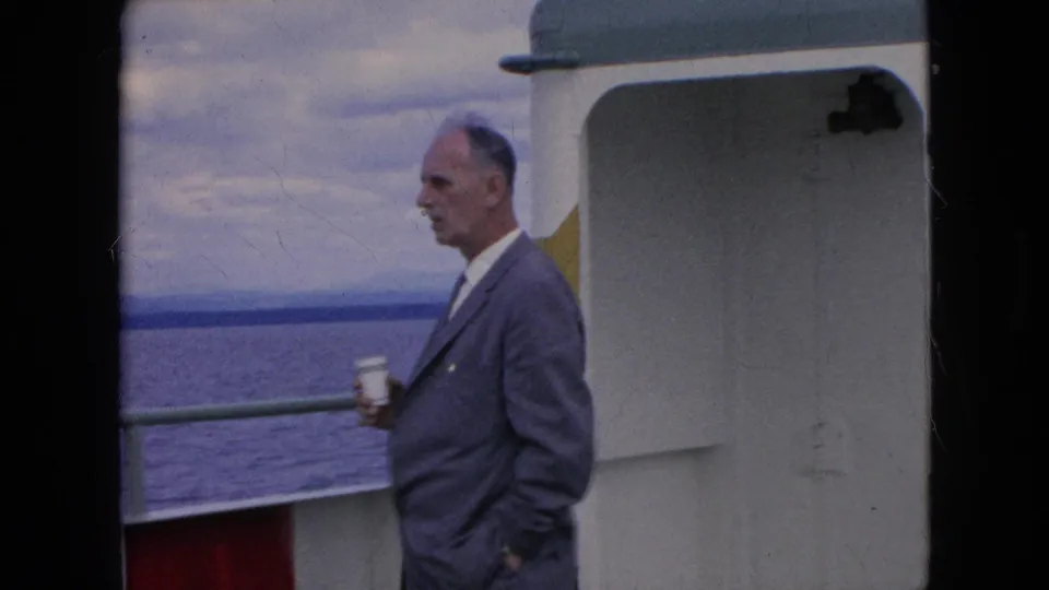 The older man remains on the ferry deck holding a cup near the white cabin wall, with open water and a cloudy horizon visible beyond the railing.