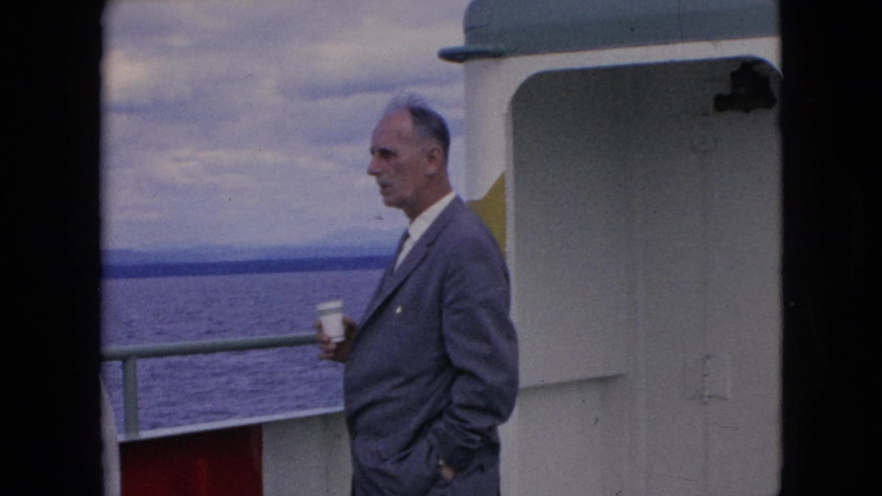 In 1964 home-movie footage, an adult man in a gray suit stands in profile on a boat deck holding a white paper cup, with railings, open water, and cloudy sky behind him.