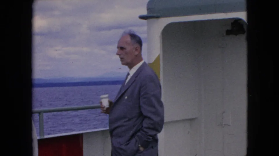 In 1964 home-movie footage, an adult man in a gray suit stands in profile on a boat deck holding a white paper cup, with railings, open water, and cloudy sky behind him.
