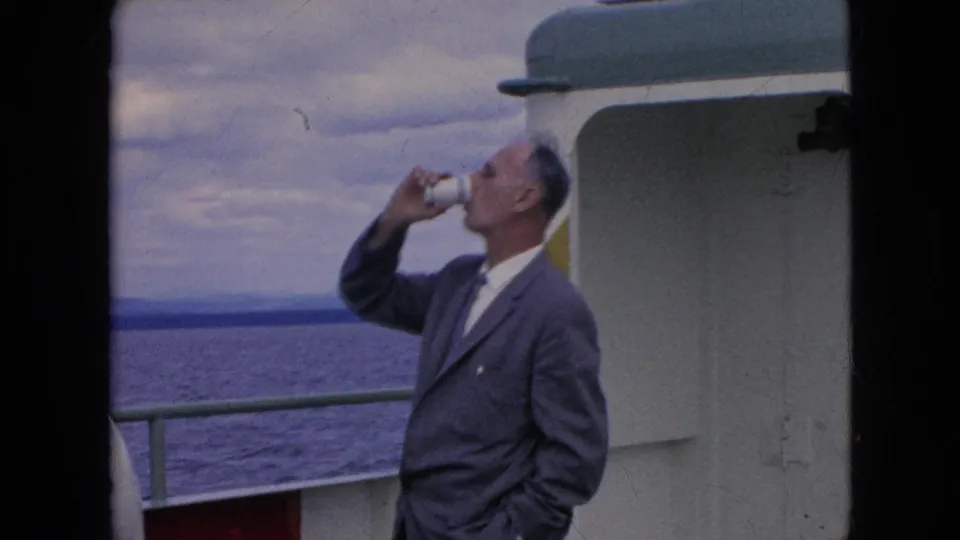 In 1964 home-movie footage, the man in a gray suit continues taking a drink from a white paper cup while standing by the railing on a boat deck.
