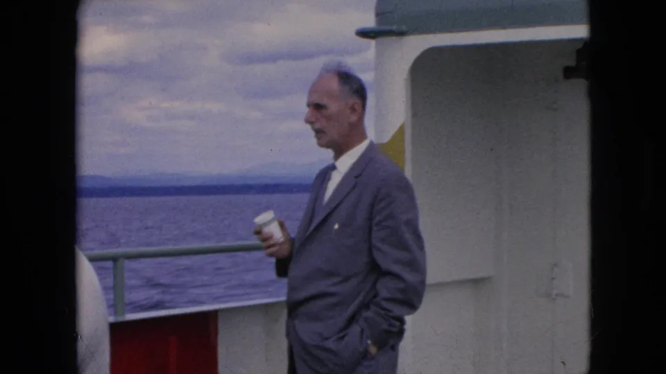 In 1964 home-movie footage, the suited man stands on the boat deck after drinking, holding the white paper cup with open water and cloudy sky behind him.