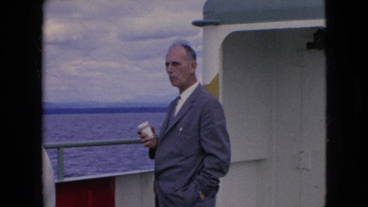 A man in a gray suit and tie holds a white cup on the deck of a moving boat, with open water, railing, cabin wall, and cloudy sky visible in 1964 travel footage.