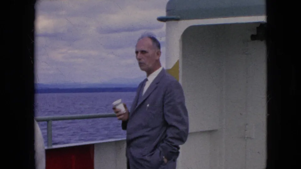 A man in a gray suit and tie holds a white cup on the deck of a moving boat, with open water, railing, cabin wall, and cloudy sky visible in 1964 travel footage.