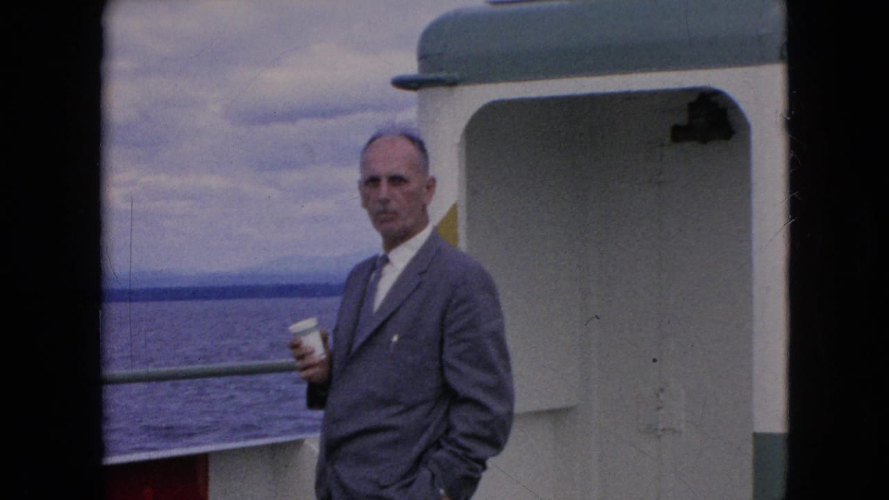Man in gray suit faces the camera while holding a cup on a boat.