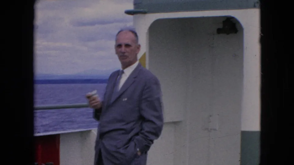 A gray-suited man stands close to the boat cabin wall, holding a cup as the vessel passes open water under a cloudy sky.