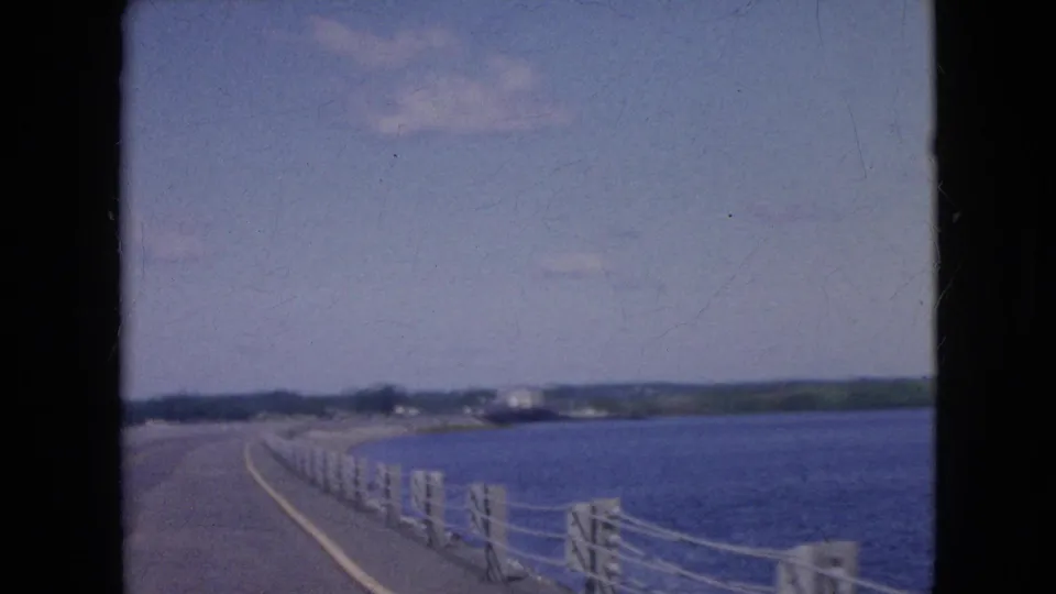 The view shifts away from the passenger to the boat railing and forward deck line, with blue water, distant shoreline, low hills, and sky visible.