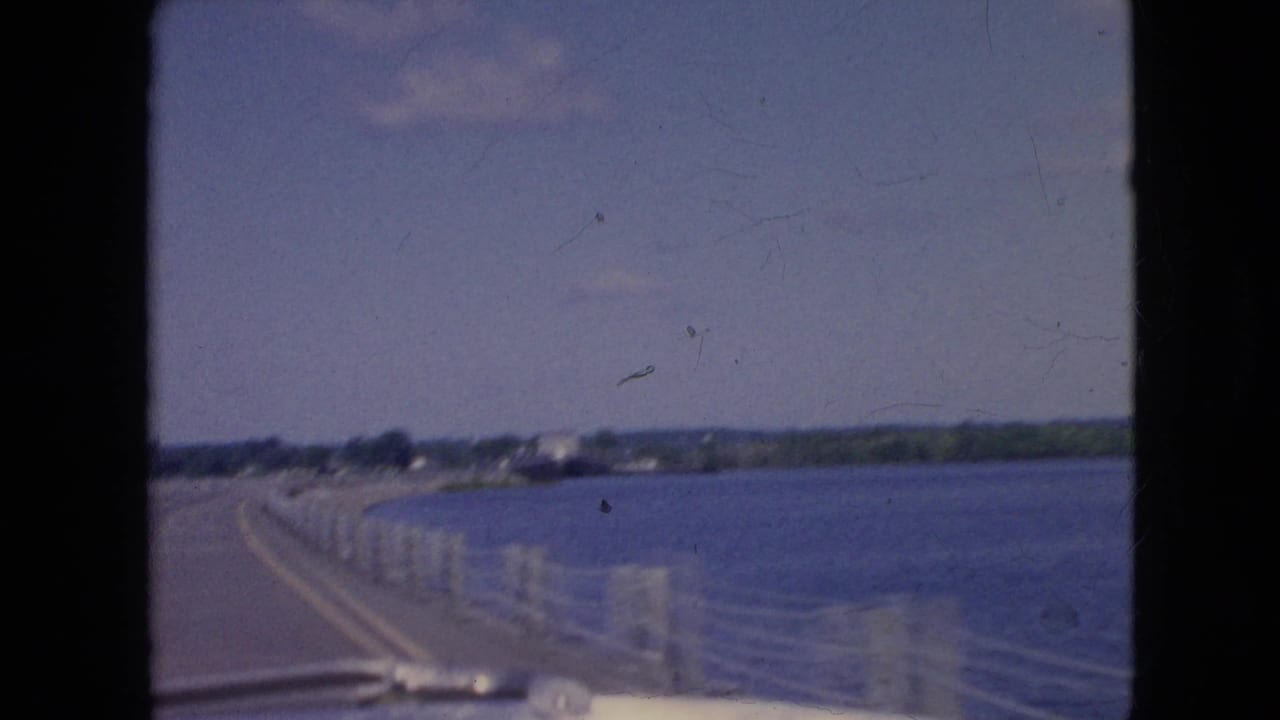 1964 8mm home-movie frame from inside a vehicle crossing a bridge near Niagara Falls, New York, with water to the right, low shoreline ahead, and pale railing posts along the road.
