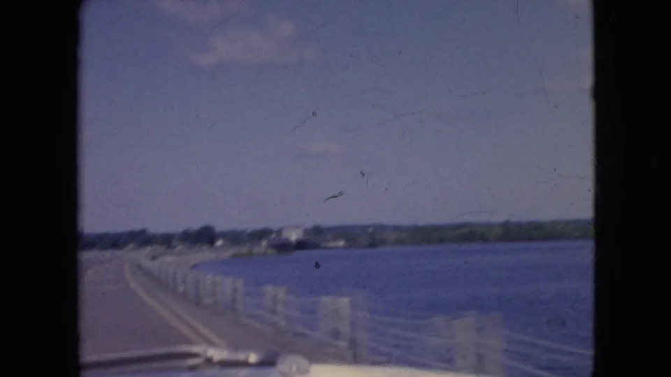 1964 8mm home-movie frame from inside a vehicle crossing a bridge near Niagara Falls, New York, with water to the right, low shoreline ahead, and pale railing posts along the road.