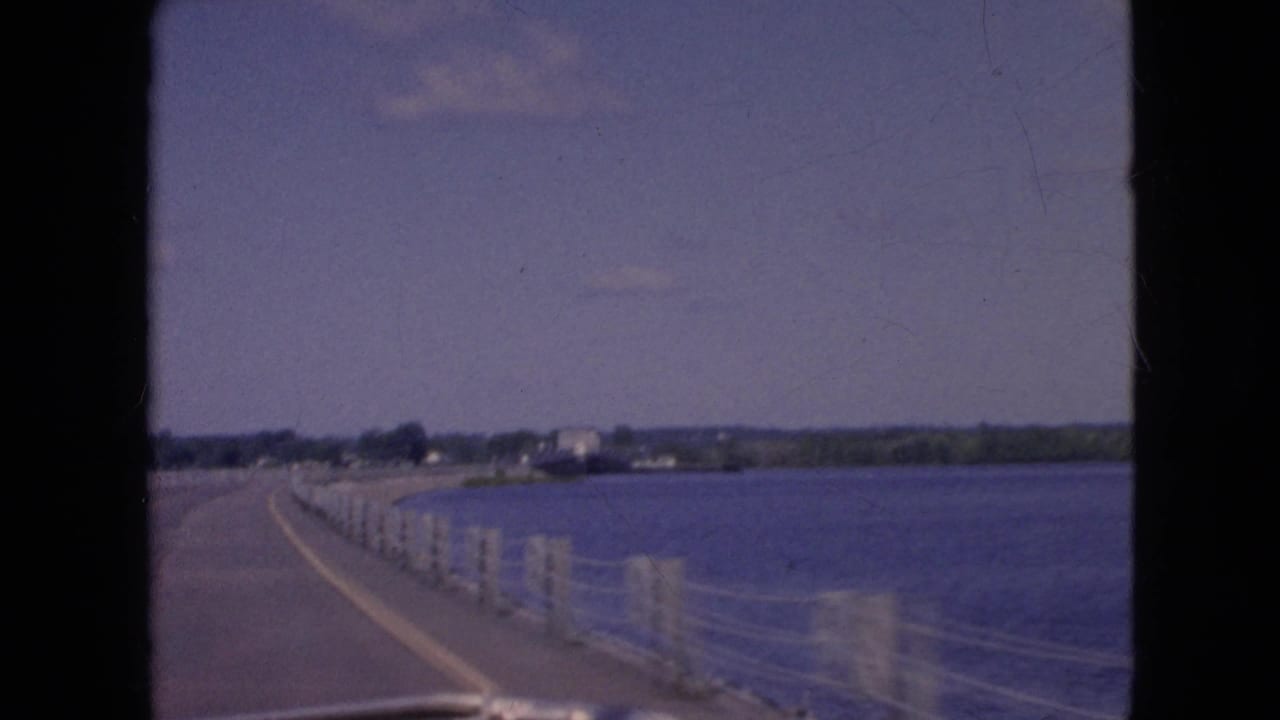 Bridge roadway and railing posts seen from inside a moving car beside open water.
