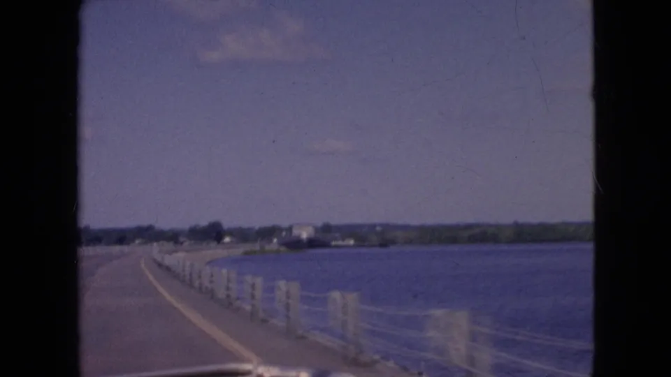 The car continues along a long bridge over water, with the dashboard edge at the bottom of the 8mm frame and a low developed shoreline visible in the distance.