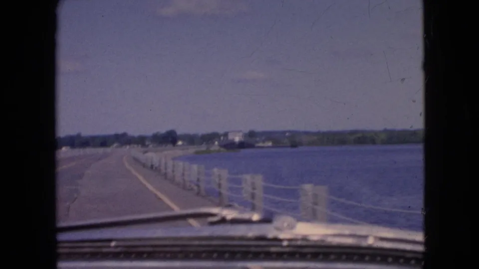 From inside the vehicle, the 8mm frame shows a curving bridge deck, dashboard trim, repeated pale posts, blue water, and a distant tree-lined shoreline near Niagara Falls, New York.