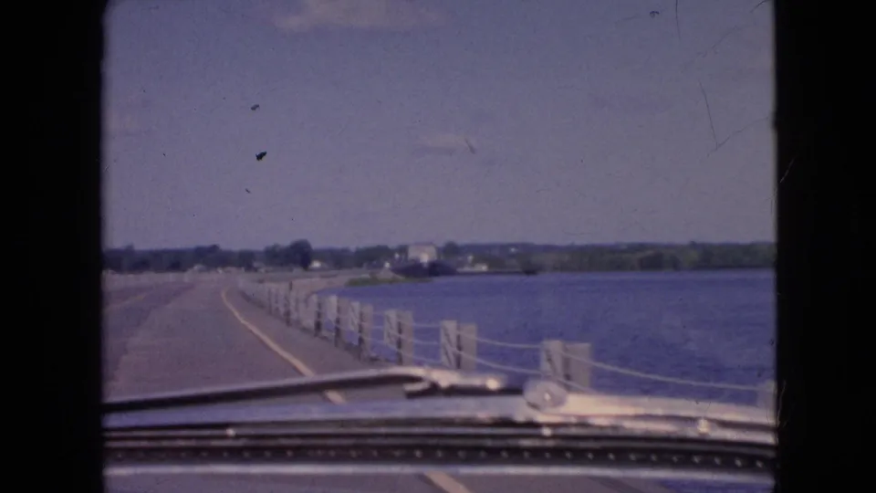 A later frame shows more of the dashboard and windshield base as the vehicle follows the bridge toward a distant shoreline, with water and pale railing posts filling the right side.