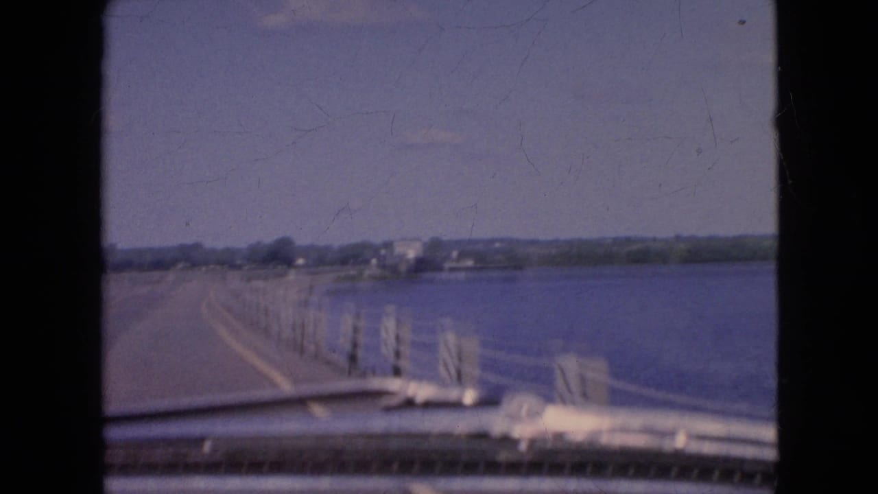 1964 8mm home-movie frame from the Niagara Falls, New York area showing a car traveling beside a broad body of water with roadside posts, cable railing, and distant buildings.