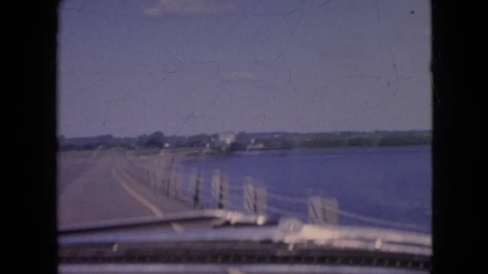 1964 8mm home-movie frame from the Niagara Falls, New York area showing a car traveling beside a broad body of water with roadside posts, cable railing, and distant buildings.