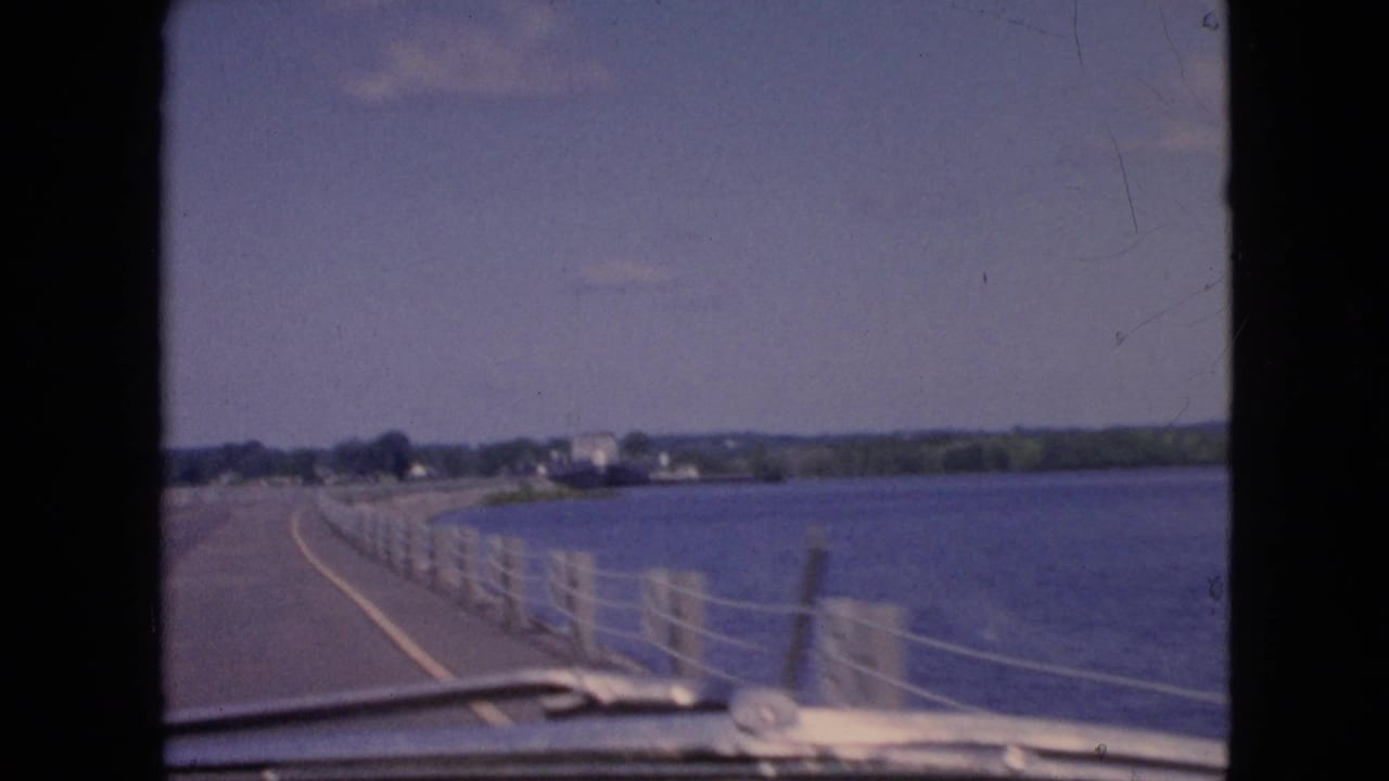 1964 Niagara Falls-area travel footage frame showing a curving road beside open water, seen from inside or just behind the front of a moving car.