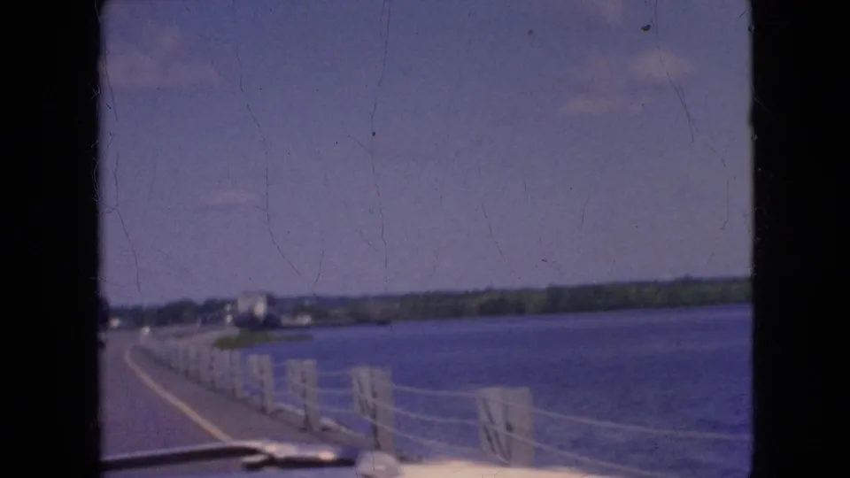 Soft-focus 1964 8mm frame showing a moving-car view of a waterside roadway with cable railing, blue water, and a low tree-lined shoreline in the distance.
