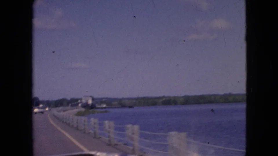 1964 Niagara Falls, New York area frame showing the end of a short moving-car shot along a waterfront road with posts, wire railing, open water, and distant shoreline houses.