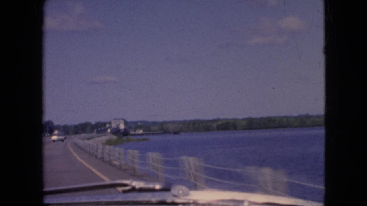 1964 8mm home-movie view from inside a moving vehicle on a river bridge near Niagara Falls, New York, with railing, water, distant cars, and shoreline visible.