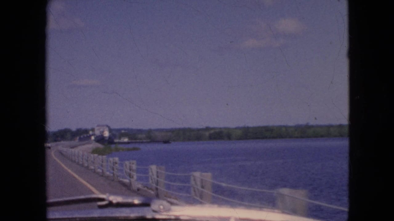 Bridge railing and river seen from a moving car with distant vehicles ahead.