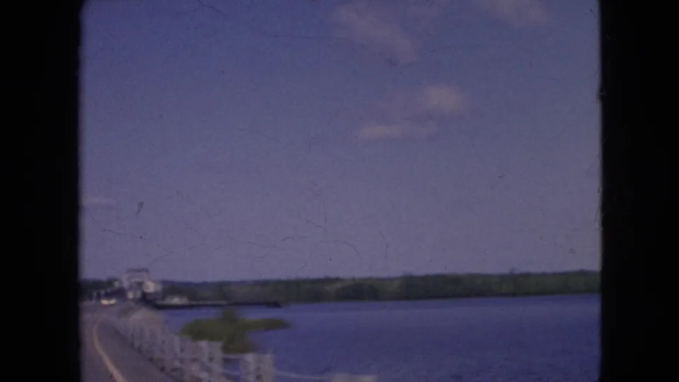 Soft 8mm frame from a moving vehicle shows the bridge railing, road edge, broad river, small green landform, distant shoreline, and open sky near Niagara Falls, New York.