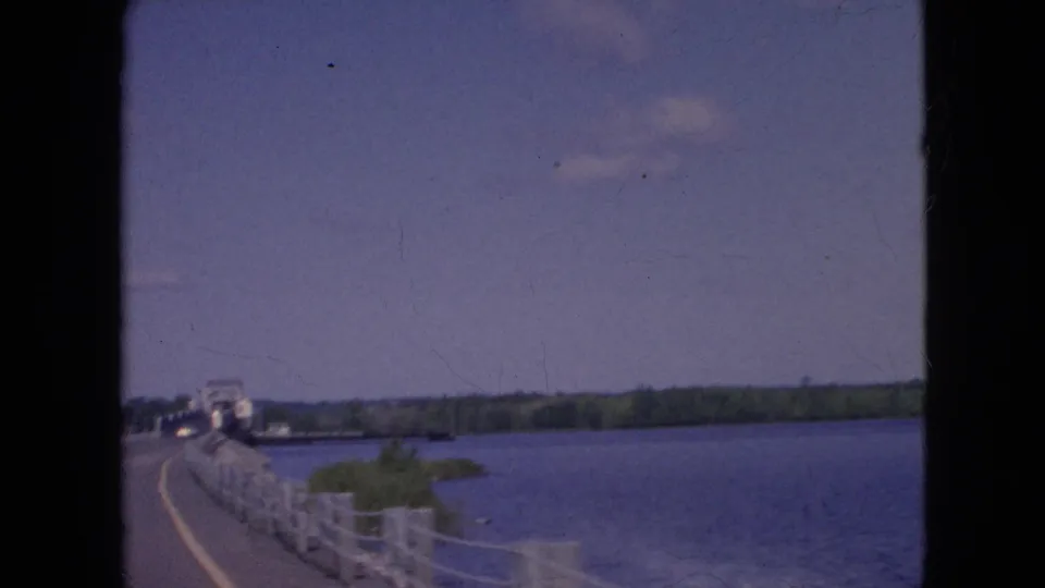 1964 home-movie frame shows a curving road and bridge railing beside a wide river, with distant vehicles and shoreline visible from inside a moving car near Niagara Falls, New York.