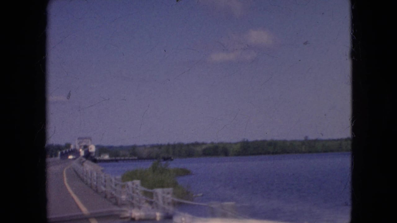 1964 8mm home-movie frame from near Niagara Falls, New York, showing a moving-car view along a bridge with white railings, blue water, and a tree-lined far shore.