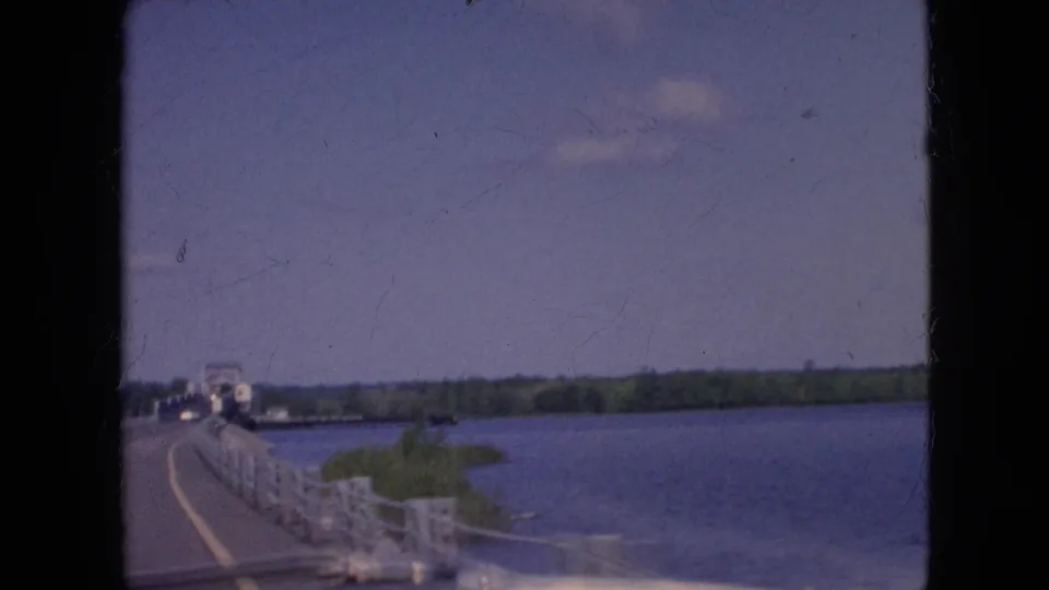 1964 8mm home-movie frame from near Niagara Falls, New York, showing a moving-car view along a bridge with white railings, blue water, and a tree-lined far shore.