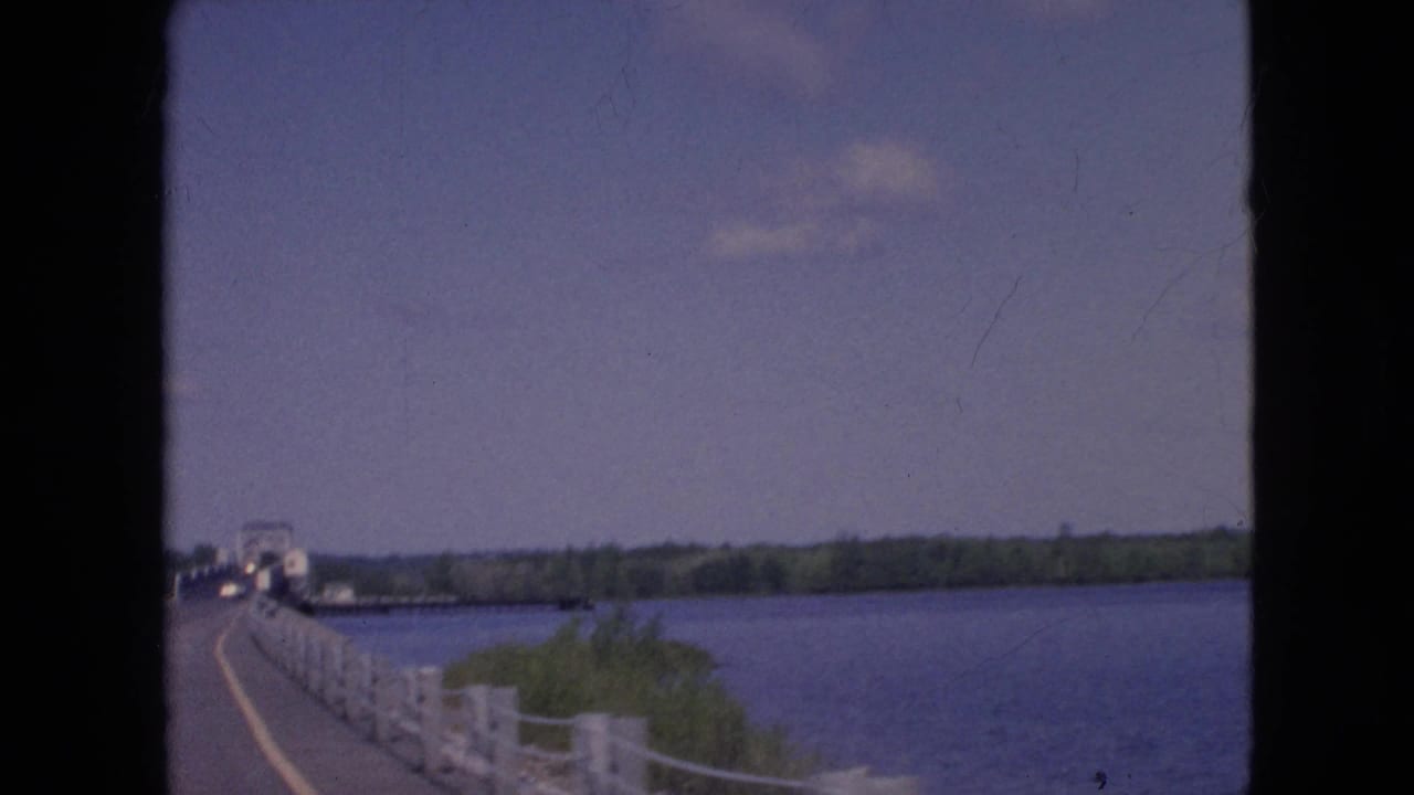 1964 visitor-shot 8mm frame near Niagara Falls, New York, looking forward along a bridge crossing with water to the right and low trees across the horizon.