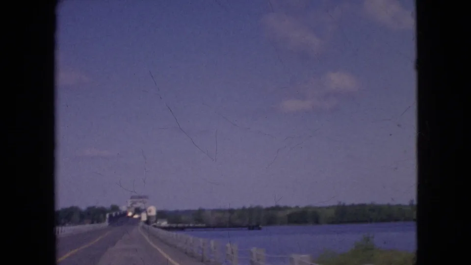 Soft 8mm frame from a 1964 Niagara-area drive showing the road cresting along a bridge, white guardrails, blue water, and a distant tree-lined shore.