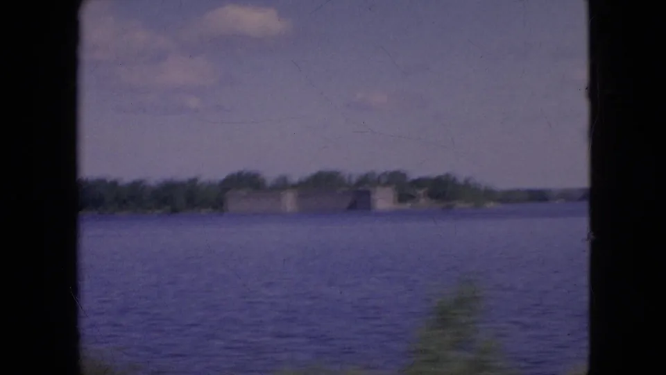 1964 8mm home-movie frame near Niagara Falls, New York, with the camera turned across open water toward a low distant bridge or shoreline structure and trees.