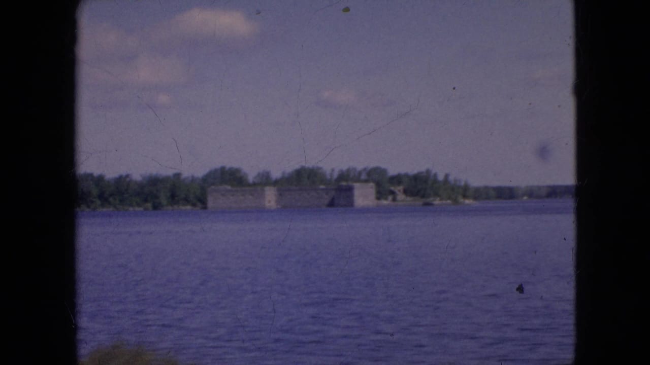 1964 8mm home-movie frame from the Niagara Falls, New York area showing a distant historic stone fort-like structure across a wide river with trees behind it.