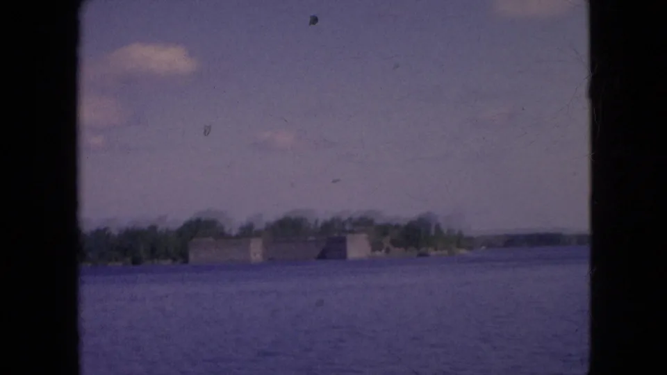 Slightly softer 1964 home-movie frame with blue-purple river water in front of a distant historic fort-like structure and wooded shoreline.