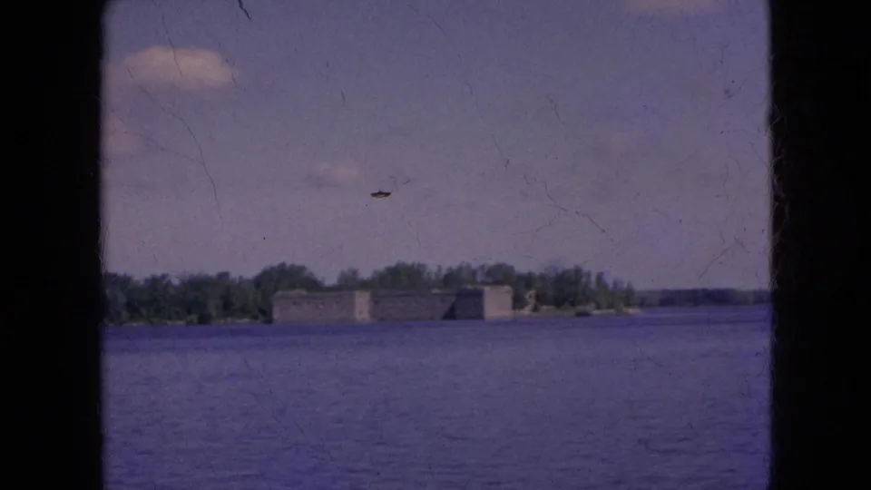 Final 1964 8mm frame showing a distant stone fort-like landmark across the river, bordered by trees and visible film scratches in the sky.