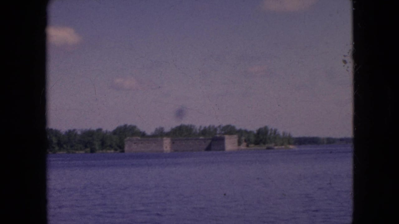 Distant waterfront view in the supplied Niagara Falls, New York clip, with a low stone building, trees, open water, and small clouds in the sky.