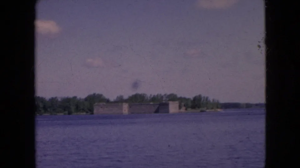 Distant waterfront view in the supplied Niagara Falls, New York clip, with a low stone building, trees, open water, and small clouds in the sky.