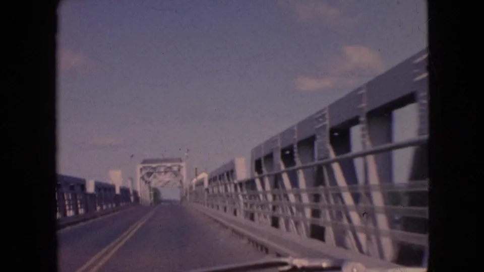 Vehicle-perspective 1964 home-movie frame on a two-lane bridge, with metal side railings, road markings, and a steel truss structure ahead.
